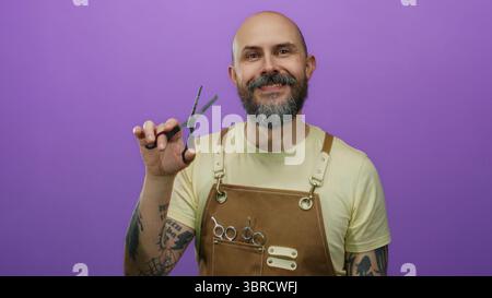 Homme barbu chauve portant un tablier et tenant des ciseaux sourit en toute confiance sur un fond violet vif mettant en vedette des bras tatoués et la coiffure t Banque D'Images