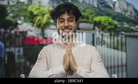 Jeune homme hispanique barbu dans des lunettes souriant avec les mains ensemble dans un geste de prière à l'extérieur entouré de verdure et de montagnes Banque D'Images