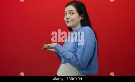 Femme en pull bleu gestes créatifs sur fond rouge vif, exprimant l'élégance et l'émotion avec des cheveux bruns et une position confiante, ble Banque D'Images