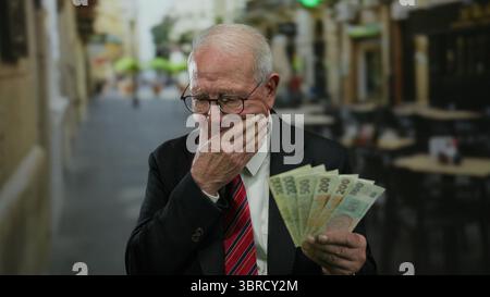Homme senior dans un costume d'affaires réfléchit tout en tenant des billets de banque de couronnes tchèques sur une terrasse de restaurant en plein air dans la rue. Banque D'Images