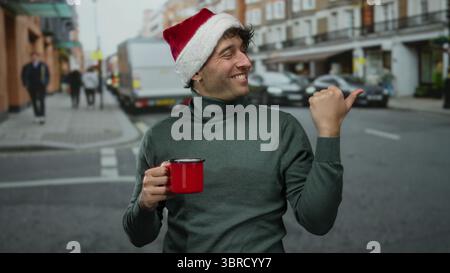 Homme hispanique portant un chapeau de noël tient une tasse rouge, souriant avec confiance dans une rue animée, avec des piétons flous et des voitures en arrière-plan, showc Banque D'Images