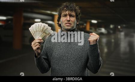 Homme hispanique tenant des roubles russes avec une expression déterminée à l'intérieur d'un garage de stationnement faiblement éclairé. Banque D'Images