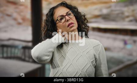 Femme mature avec des cheveux bouclés et des lunettes sur une plage touchant son cou, portant un pull, montrant une expression sereine, en plein air au bord de la mer avec bl Banque D'Images