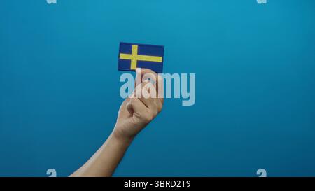 Homme tenant le drapeau suédois brodé sur fond bleu isolé, mettant en valeur l'identité nationale avec l'accent sur les détails de la main et les riches symboli culturels Banque D'Images