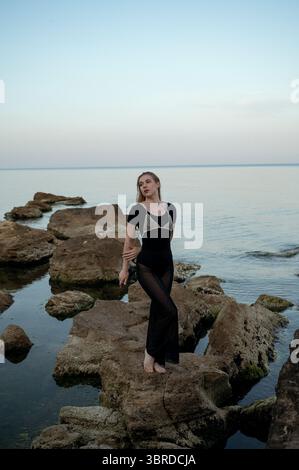 jeune femme, blonde pose sur les rochers sur fond de mer du soir Banque D'Images