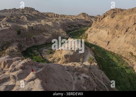 Vue aérienne d'une figure solitaire dans un rouge vif debout au sommet des falaises accidentées et ensoleillées surplombant la vallée luxuriante et verdoyante en contrebas, Kerman, Kerman Provin Banque D'Images
