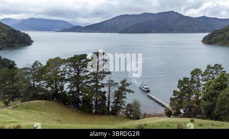 Vue aérienne d'un bateau arrivant à un quai de Wilson Bay entouré de collines verdoyantes et d'eaux calmes reflétant le ciel couvert, Marlborough Sounds, New Banque D'Images