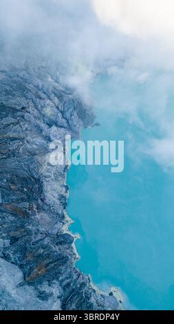Vue aérienne du lac turquoise niché dans le cratère, bordé de falaises escarpées entourées de brume éthérée, cratère du volcan Ijen, Java oriental, Indonésie. Banque D'Images