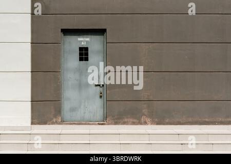 Une porte industrielle en métal gris avec une fenêtre barrée et un cadenas, située dans un mur à panneaux brun foncé avec des marches blanches. ÉTIQUETÉ « PANNEAU RUANG A-1 ». Banque D'Images