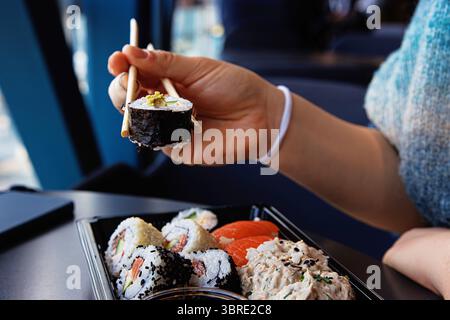 Gros plan de la main de la personne ramassant un rouleau de sushi avec des baguettes sur un plateau noir dans un café moderne. Mise au point sélective Banque D'Images