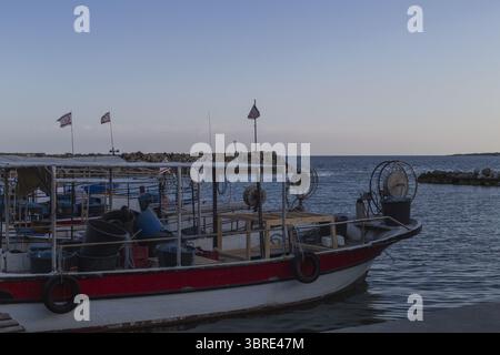 Vue sur un bateau de pêche rouge et blanc se trouve paisiblement dans le port, reflétant la douce lumière du crépuscule sur les eaux calmes, Dipkarpaz, Chypre du Nord, Cipro. Banque D'Images