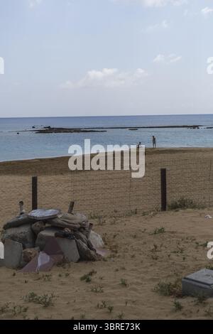 Vue sur le sable ensoleillé rencontre les eaux turquoises, une vue côtière sereine avec des rochers dispersés et des personnages lointains, Varosha, Famagouste, Chypre. Banque D'Images