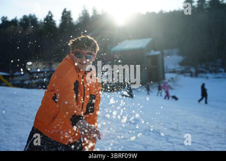 Adolescent en veste orange jetant joyeusement de la neige dans un paysage d'hiver. L'image capture un moment ludique avec la lumière du soleil et la forêt en arrière-plan, salut Banque D'Images