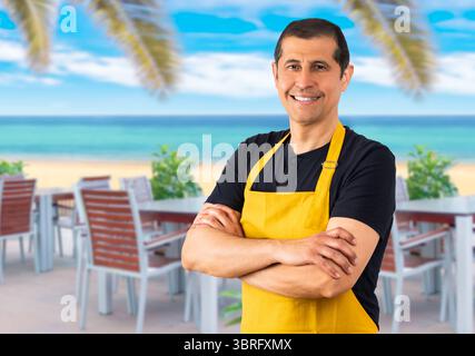 Bel homme portant employé avec tablier à l'hôtel de plage.souriant dentiellement avec les bras croisés exprimant, émotions positives Banque D'Images