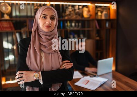 Femme d'affaires confiante portant un hijab debout avec les bras croisés dans un bureau moderne, un collègue masculin travaille sur un ordinateur portable en arrière-plan Banque D'Images