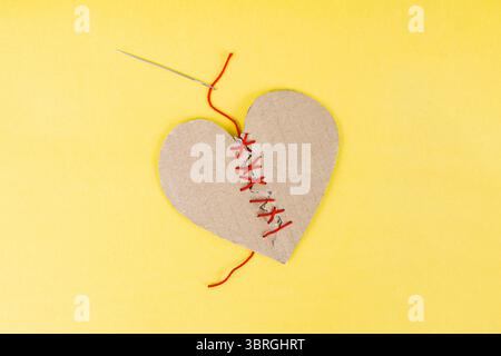 Coeur en carton cassé cousu avec une aiguille et du fil rouge sur fond jaune, conseil matrimonial Banque D'Images