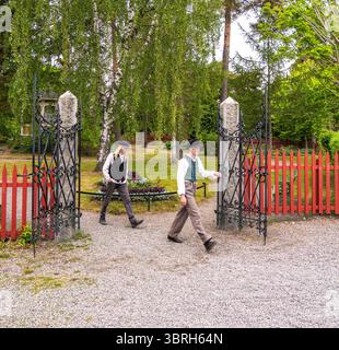 Sodertalje, Suède - 6 juin 2025 : deux jeunes hommes en tenue traditionnelle suédoise marchant à travers une porte ornée dans un jardin, entouré de verdure Banque D'Images