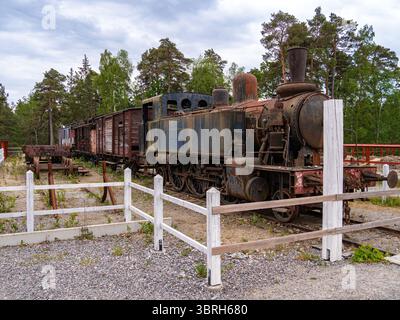 Sodertalje, Suède - 6 juin 2025 : une vieille locomotive à vapeur et des wagons de marchandises d'époque stationnés sur une piste de gravier, entourés d'arbres et d'une clôture blanche. T Banque D'Images