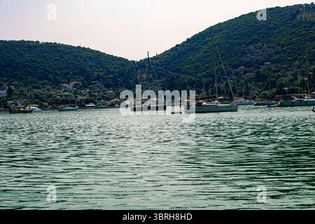 Voiliers ancrés dans des eaux vertes calmes près d'un village côtier avec des collines boisées, paysage estival typiquement méditerranéen. Banque D'Images