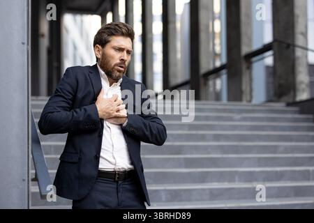 Un homme d'affaires inquiet en costume agrafe sa poitrine tout en se tenant debout à l'extérieur d'un bâtiment, possiblement éprouvant des problèmes de santé ou de stress. Banque D'Images