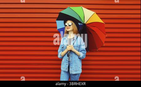 Portrait d'automne de jeune femme élégante et souriante tenant un parapluie coloré portant une veste de jean sur fond rouge Banque D'Images