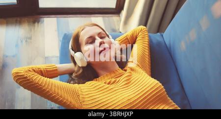 Heureuse jeune femme détendue écoutant de la musique avec des écouteurs sans fil avec les yeux fermés se trouve sur le canapé à la maison Banque D'Images