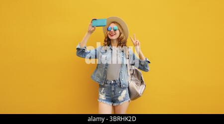 Portrait de jeune femme élégante et souriante prenant selfie avec téléphone et sac à dos en chapeau de paille touristique d'été, vêtements en denim sur dos jaune vif Banque D'Images