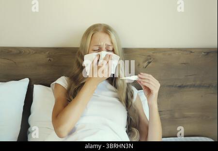 Malade bouleversée femme éternuant nez de soufflage à l'aide de mouchoir en papier et en mesurant la température corporelle en regardant le thermomètre sur le lit dans la chambre à coucher à la maison Banque D'Images