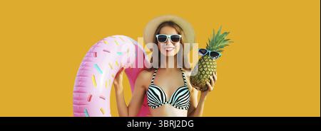 Vacances d'été, tourisme, jeune femme souriante heureuse avec anneau gonflable de natation et ananas portant chapeau de paille sur fond jaune de studio Banque D'Images
