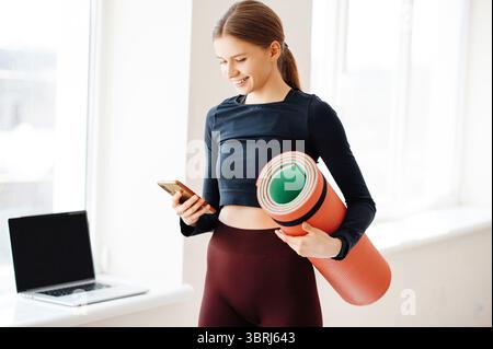 Belle jeune blonde dans des vêtements de sport debout près de la fenêtre avec ordinateur portable, tenant le tapis de fitness à la main et lisant le message sur smartphone. Dame souriante Banque D'Images
