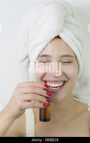 Heureuse jeune femme avec une serviette sur la tête tenant une bouteille en verre ambré de sérum de peau. Belle jeune femme brésilienne hydrate sa peau avec isolat de sérum Banque D'Images