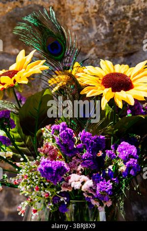 Cette image présente un bouquet vibrant de tournesols artificiels, de fleurs violettes et de plumes de paon disposées dans un vase transparent. Banque D'Images