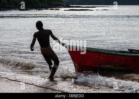 un pêcheur local tire son bateau sur une plage de sable en silhouette en fin d'après-midi sur l'île de sao tome alors que les vagues chevauchent le rivage Banque D'Images
