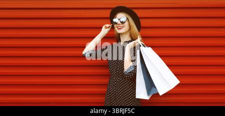 Portrait de jeune femme souriante heureuse avec des sacs à provisions portant un chapeau rond noir sur fond rouge Banque D'Images