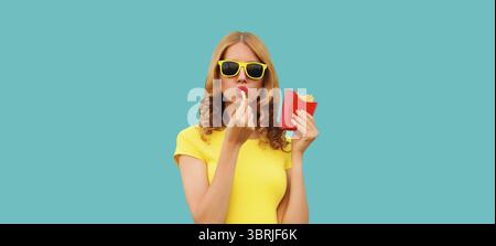 Portrait de jeune femme élégante mangeant des frites, de la nourriture rapide de pommes de terre frites dans des lunettes de soleil sur fond bleu de studio Banque D'Images