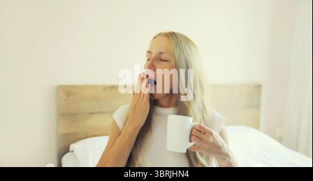 Paresseuse femme d'âge moyen se réveillant après avoir dormi et bâillant sur le lit tient une tasse de café dans la chambre à coucher à la maison Banque D'Images