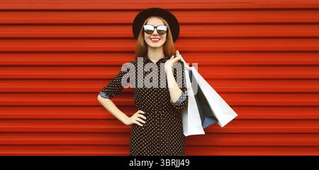 Portrait de jeune femme souriante heureuse avec des sacs à provisions portant un chapeau rond noir sur fond rouge Banque D'Images