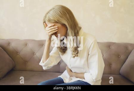 Jeune femme a mis les mains sur l'abdomen souffrant de maux d'estomac saisissant chellyache se sentir mal avec douleur assis sur un canapé à la maison Banque D'Images