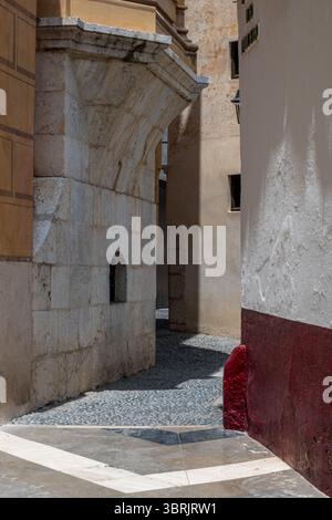 Une ruelle étroite entre des bâtiments en pierre baignés de soleil, révélant les textures anciennes de l’architecture historique de Málaga. Banque D'Images