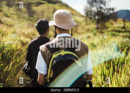 Deux hommes d'Asie du Sud-est marchant sur un étroit sentier de montagne à travers de hautes herbes sous la lumière du jour, portant des vêtements décontractés et des sacs à dos. Banque D'Images