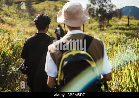 Deux hommes d'Asie du Sud-est marchant sur un étroit sentier de montagne à travers de hautes herbes sous la lumière du jour, portant des vêtements décontractés et des sacs à dos. Banque D'Images