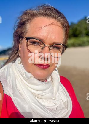 Femme avec des lunettes et une écharpe blanche pose à l'extérieur, portant une tenue rouge, avec un ciel bleu clair en arrière-plan, rayonnant de confiance et d'élégance Banque D'Images