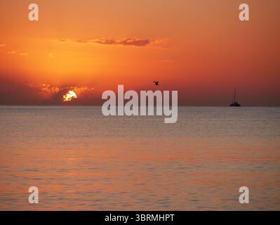 Un coucher de soleil à couper le souffle sur la mer calme avec un voilier à l'horizon. Des tons chauds orange et dorés remplissent le ciel et se reflètent sur l'eau. Voyage, na Banque D'Images