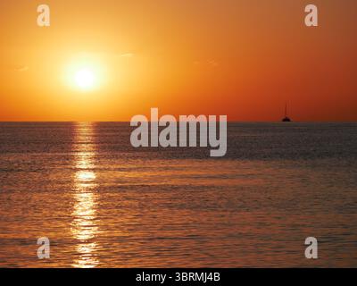 Un coucher de soleil à couper le souffle sur la mer calme avec un voilier à l'horizon. Des tons chauds orange et dorés remplissent le ciel et se reflètent sur l'eau. Voyage, na Banque D'Images