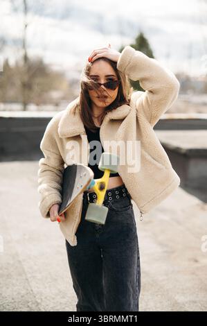 Jeune femme dans une veste beige tenant une planche à roulettes à l'extérieur. Banque D'Images