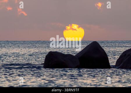 Le grand orbe jaune du soleil couchant touche l'horizon océanique derrière des rochers silhouettes à la plage d'Anse Lazio aux Seychelles. Banque D'Images