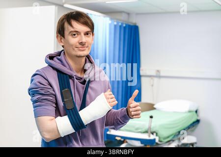 Jeune homme avec le bras cassé dans des sourires d'écharpe, levant le pouce près du lit d'hôpital vide dans la salle d'urgence, récupération de blessure, patient heureux Banque D'Images