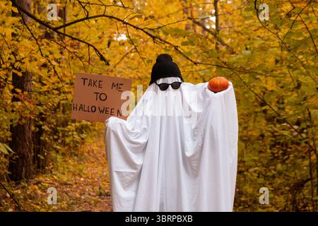 Joyeuses vacances d'Halloween. Mignon méconnaissable personne tenant le carton avec le texte ME PRENDRE À HALLOWEEN et citrouille en costume fantôme sur fond de forêt d'automne. Costume créatif avec chapeau blanc et noir et lunettes. Concept minimal Banque D'Images