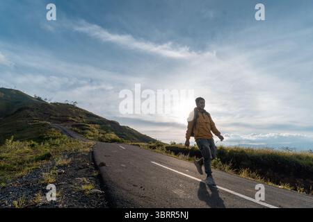 Randonneur en veste jaune tenant un téléphone tout en marchant seul sur une route vallonnée. Banque D'Images