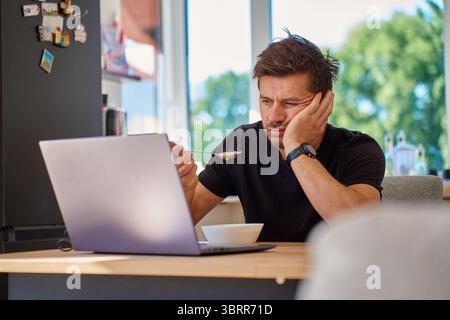 Homme assis à la table de la cuisine, mange de la bouillie d'avoine avec un regard mécontent et a poussé son assiette loin. Homme malheureux mangeant un petit déjeuner sans goût dans la cuisine Banque D'Images